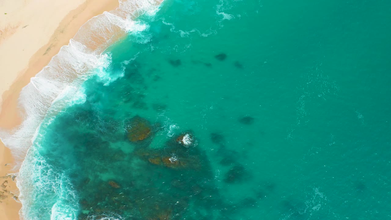 Overhead frame at Secrets Marquis Los Cabos, Mexico, showing a broader surf band along the left beach, green-blue water over dark rock patches, and no visible people.