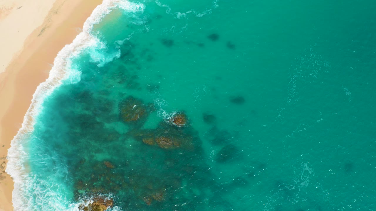 At Secrets Marquis Los Cabos, Mexico, the opening overhead frame shows pale sand on the left, white surf curling into clear green water, submerged dark rock patches, and no visible people.