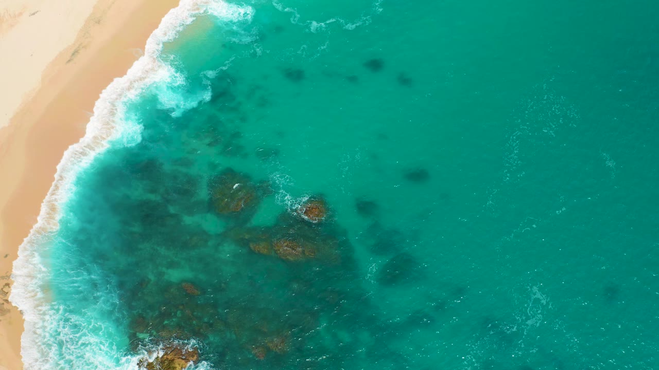 One second in, the aerial view at Secrets Marquis Los Cabos, Mexico keeps the beach along the left edge while foamy surf spreads farther over green water with no visible people.