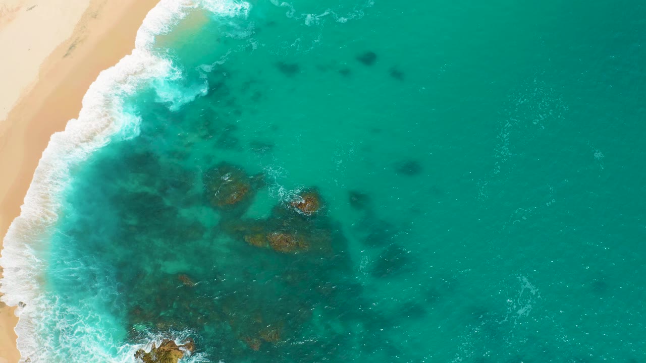 At 0:02, the Secrets Marquis Los Cabos, Mexico shoreline remains top-down, with a brighter band of foam near the sand and open turquoise water filling the right side with no visible people.