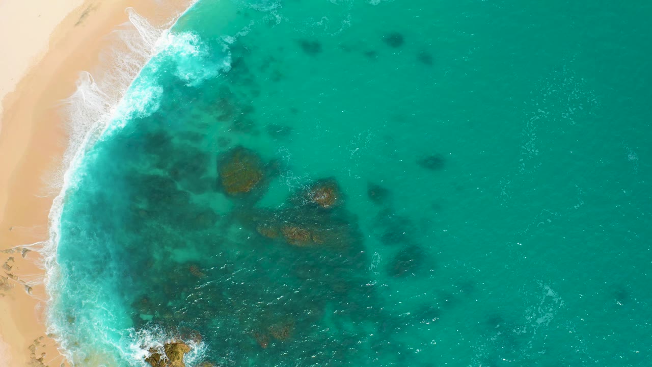 Aerial frame over Secrets Marquis Los Cabos, Mexico, showing the wave line pushed farther along the sandy edge, with dark rock patches beneath turquoise water and no visible people.