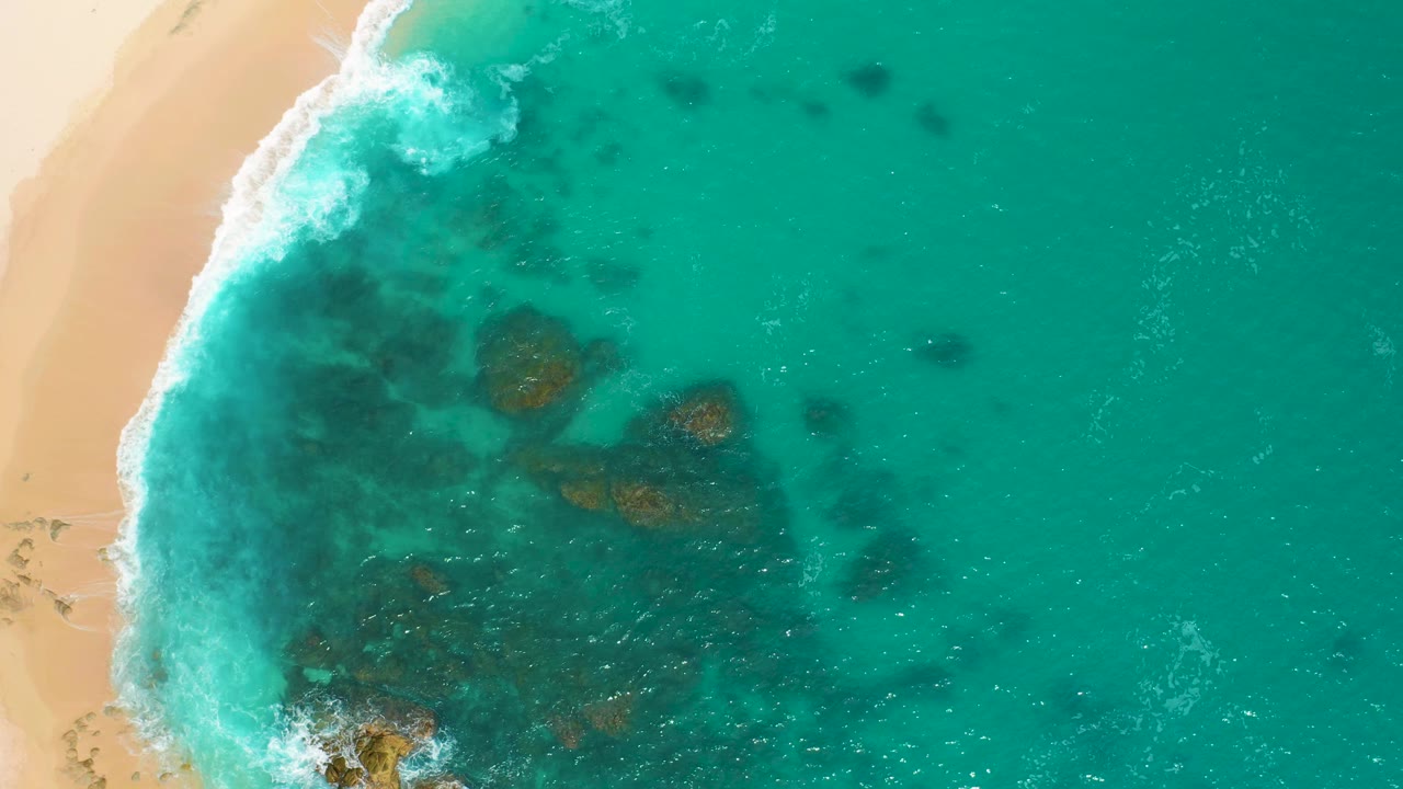 Overhead frame from Secrets Marquis Los Cabos, Mexico, with white foam breaking along the left shoreline, rocky shallows under green-blue water, and no visible people.