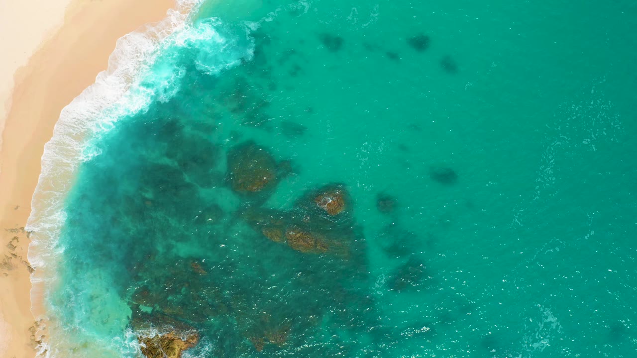 Aerial shoreline with white surf and rocks under clear blue-green water.