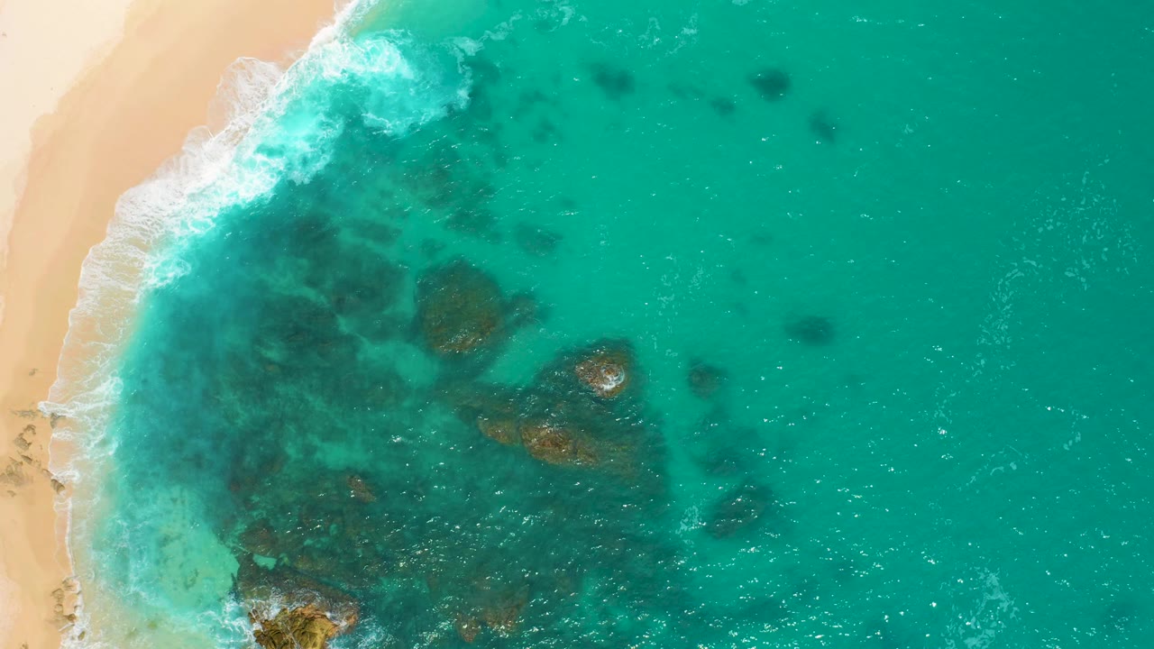 Top-down view at Secrets Marquis Los Cabos, Mexico, showing a pale beach strip on the left, a breaking foam line, scattered dark underwater patches, and no visible people.