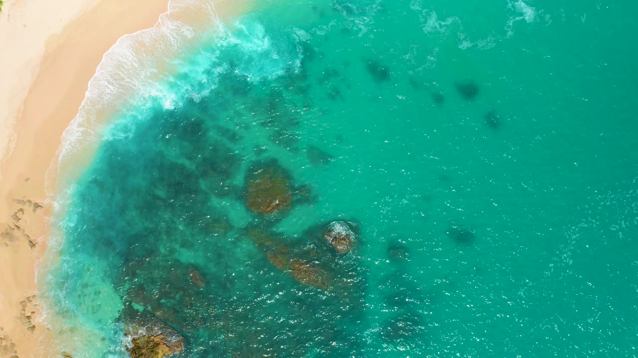 Aerial frame over Secrets Marquis Los Cabos, Mexico, showing the surf line curving along the beach, several dark underwater rock patches, bright ripples, and no visible people.