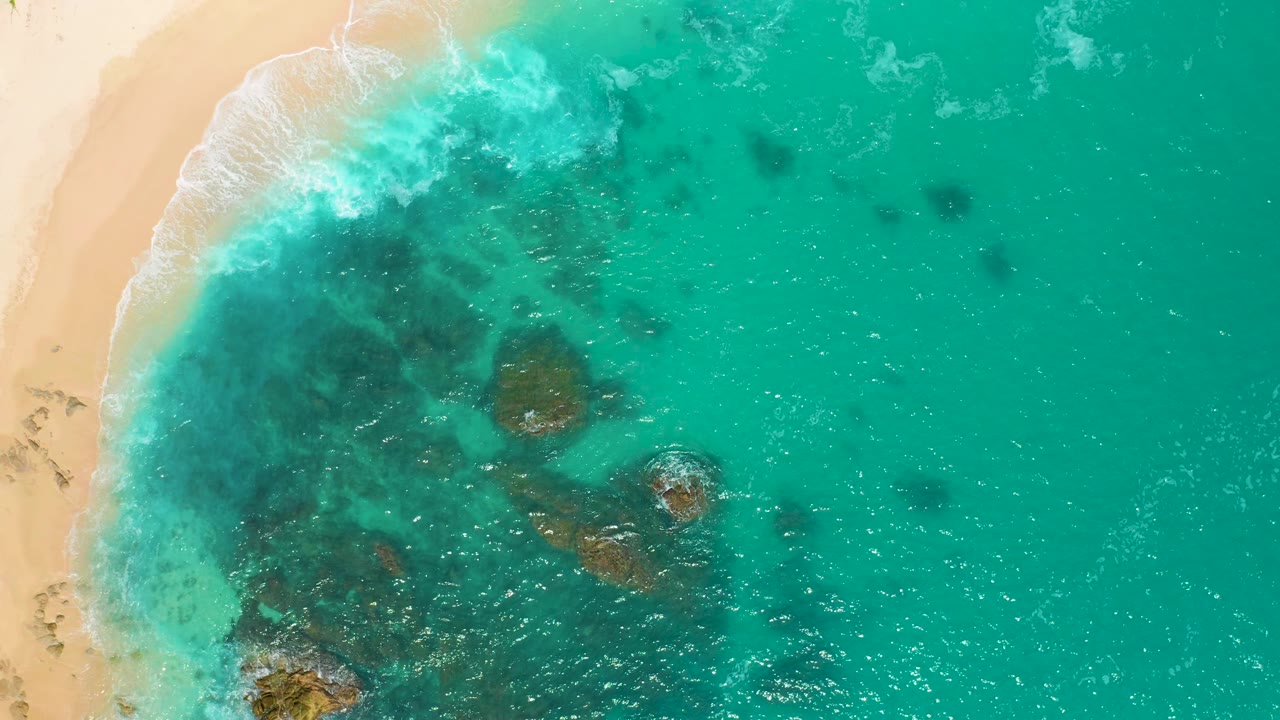 Near-vertical drone view at Secrets Marquis Los Cabos, Mexico, with tan sand on the left, aqua water across the frame, dark submerged formations, small foam trails, and no visible people.