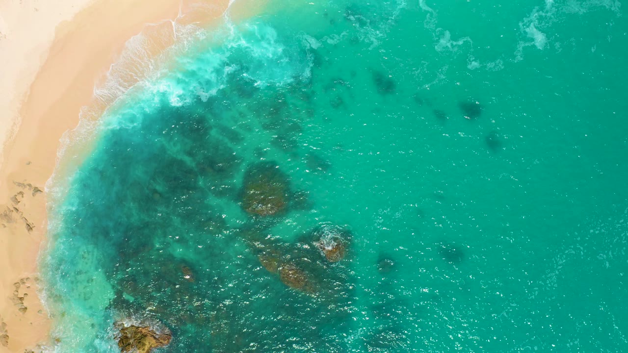 Overhead view at Secrets Marquis Los Cabos, Mexico, where pale sand and surf occupy the left side, a broad aqua water field extends right, underwater rocks sit near the lower edge, and no people are visible.