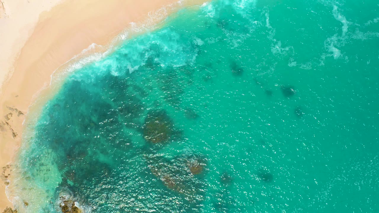 Aerial frame from Secrets Marquis Los Cabos, Mexico, with foamy surf along the sand at upper left and darker underwater patches spread through the green water, no visible people.