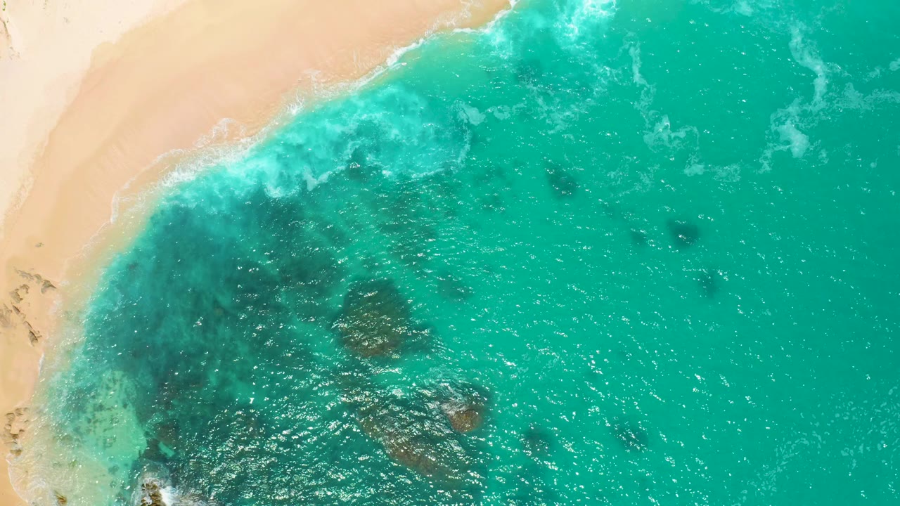 Top-down view at Secrets Marquis Los Cabos, Mexico, where small white waves reach the sand and broad turquoise water fills most of the frame, with no visible people.