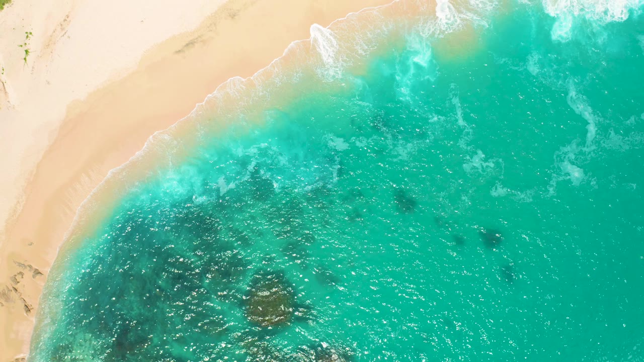 Top-down coastal frame at Secrets Marquis Los Cabos, Mexico showing a curved sand line, translucent aqua shallows, white foam at the beach, and no visible people.