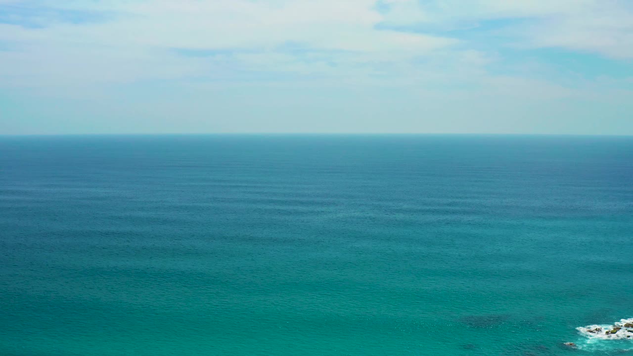 Blue-green ocean off Secrets Marquis Los Cabos, Mexico, with the horizon high in frame, faint surface bands, a rocky whitewater patch at the lower right, and no visible people.