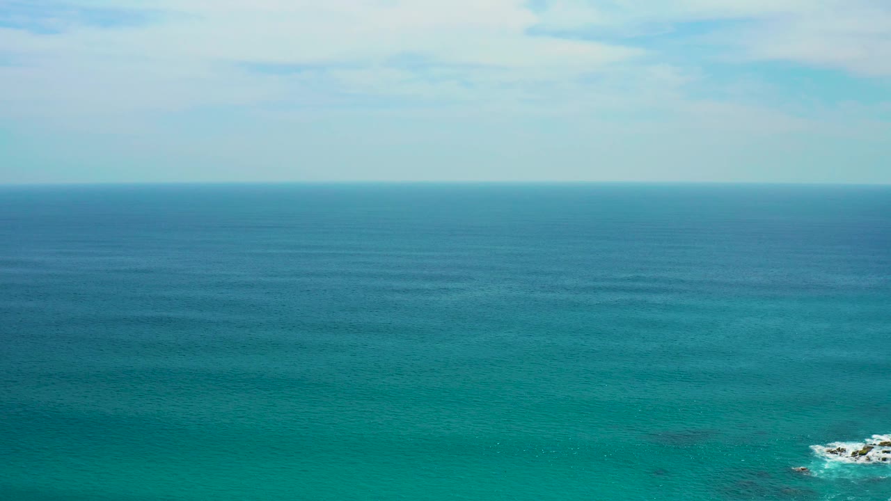 Wide turquoise ocean under a pale sky with a small rocky break near the lower left.