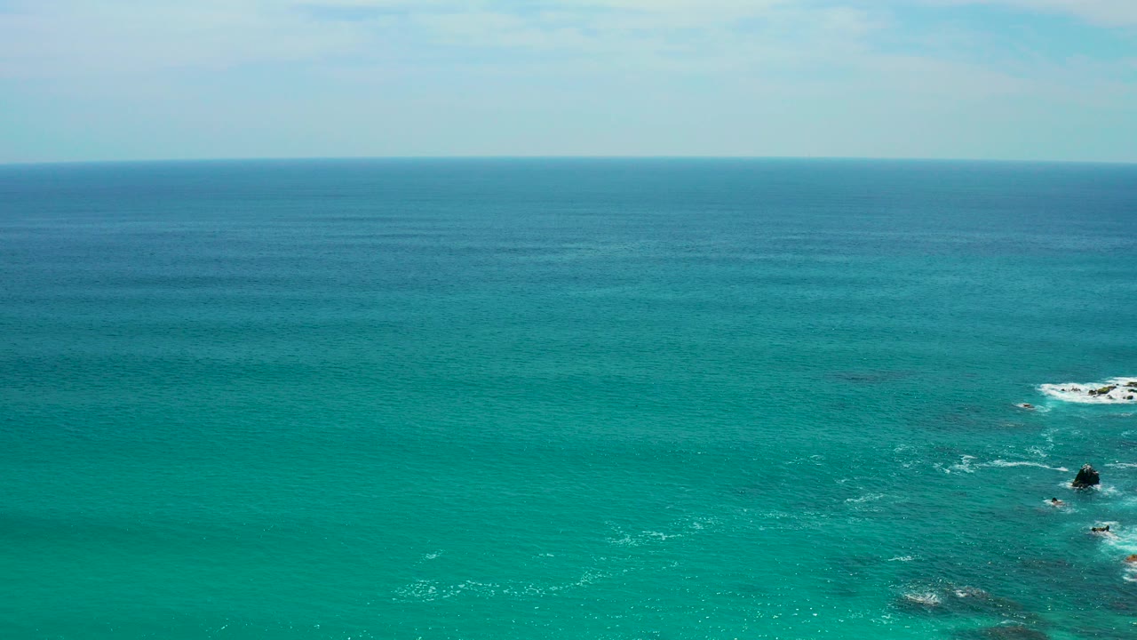 Wide aerial view over turquoise water at Secrets Marquis Los Cabos, Mexico, with a level horizon, small dark rocks near the right side, white wave foam, and no visible people.