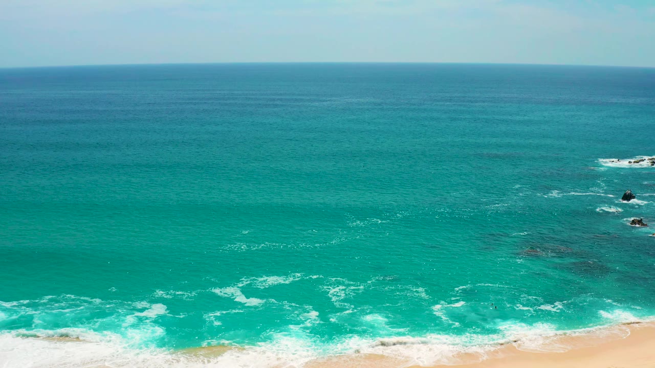 Aerial coastal frame at Secrets Marquis Los Cabos, Mexico, showing blue-green water, thin white wave lines approaching the sand, dark rock patches offshore, and no visible people.