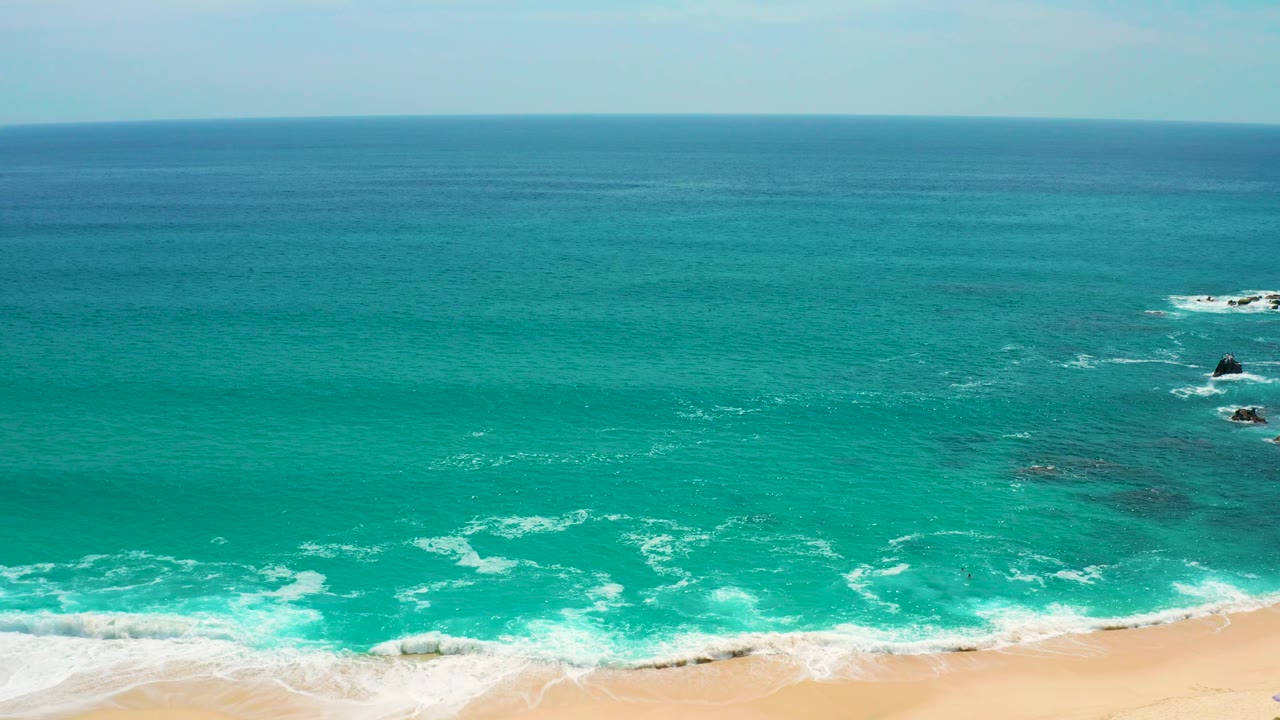 Secrets Marquis Los Cabos, Mexico beach view with the shoreline visible along the bottom, foamy surf curling over turquoise water, small offshore rocks near the right side, and no visible people.