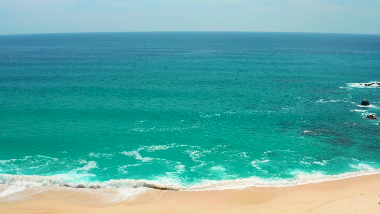 Elevated Secrets Marquis Los Cabos, Mexico shoreline frame with bright white surf washing onto sand, dark rocks breaking the water surface offshore, a broad turquoise sea, and no visible people.