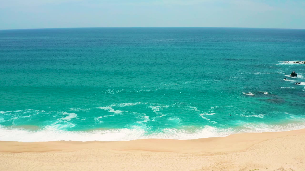 Early frame shows the Secrets Marquis Los Cabos beach from above, with blue-green water, a wider strip of foamy shore break, pale sand, and no visible people.
