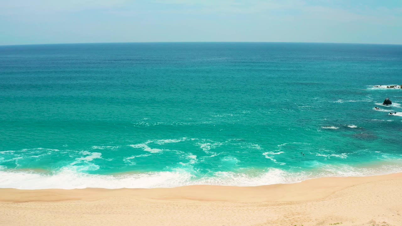 Mid-early frame holds on the Secrets Marquis Los Cabos coast with open turquoise water, white foam near the beach, dark rocks near the right edge, and no visible people.