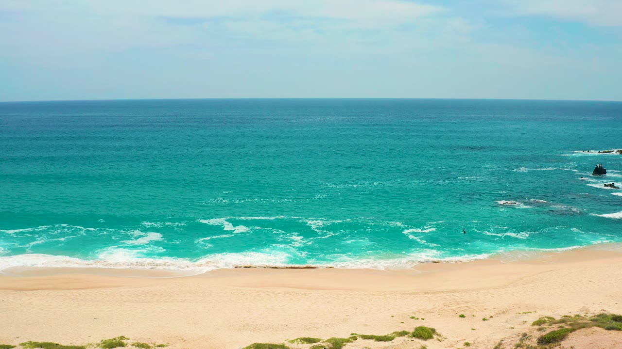 Elevated coastal frame at Secrets Marquis Los Cabos, Mexico, showing blue-green ocean bands, foamy shorebreak, pale sand, low dunes, distant rocks, and no visible people.