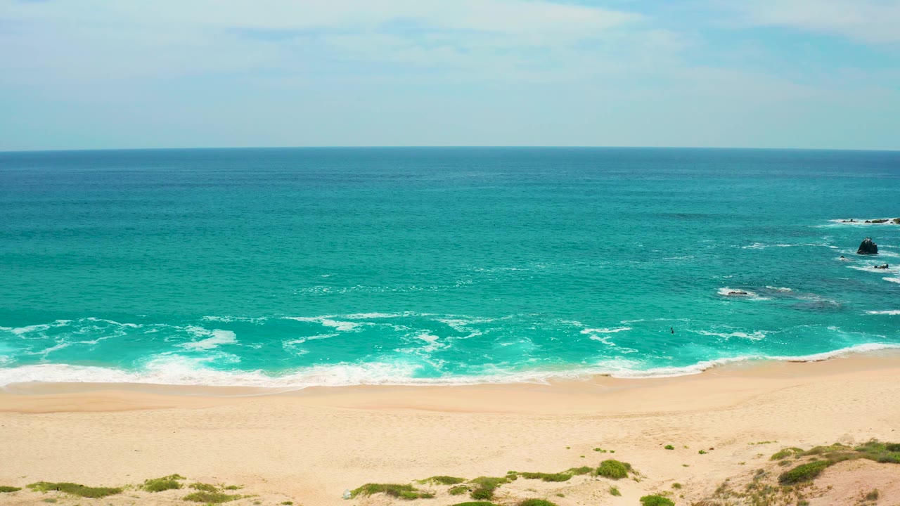 Secrets Marquis Los Cabos, Mexico coastline view with a straight horizon, teal ocean, white surf seams, wide sand, dune scrub below, offshore rock outcrops, and no visible people.
