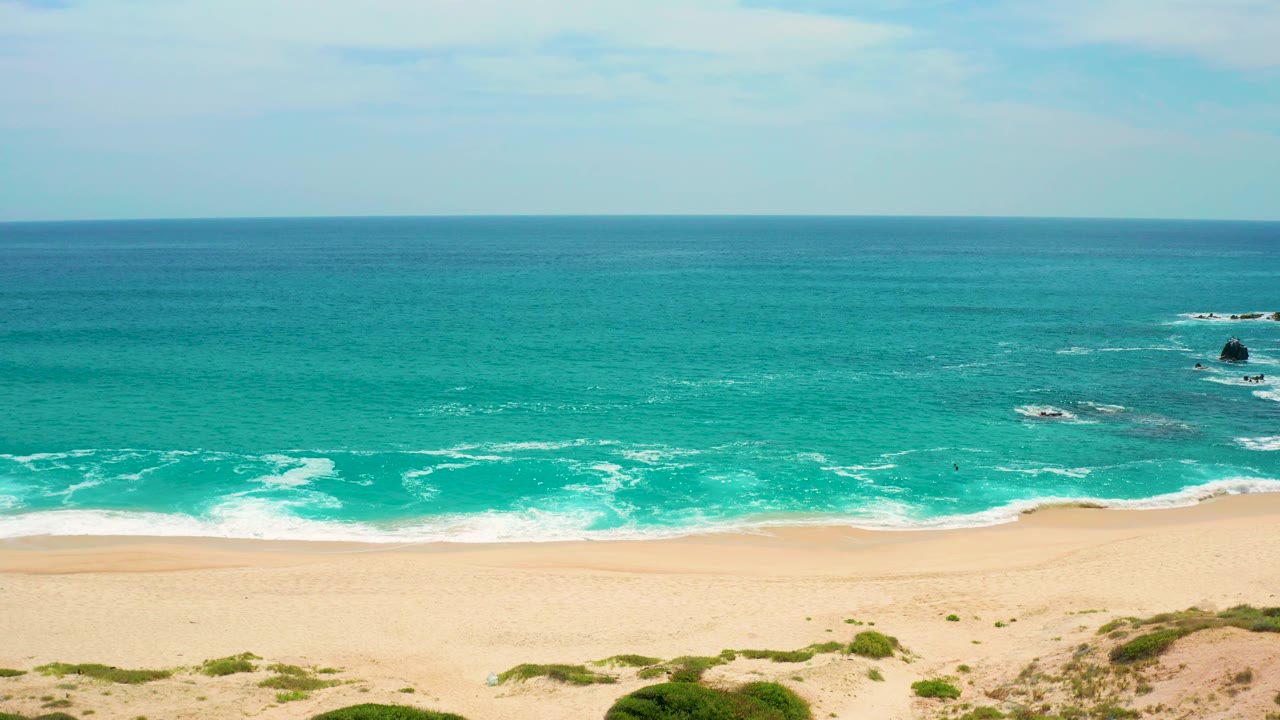 Elevated view from Secrets Marquis Los Cabos, Mexico, with foamy waves reaching a long golden beach, turquoise water beyond, low vegetation at the bottom edge, and no visible people.