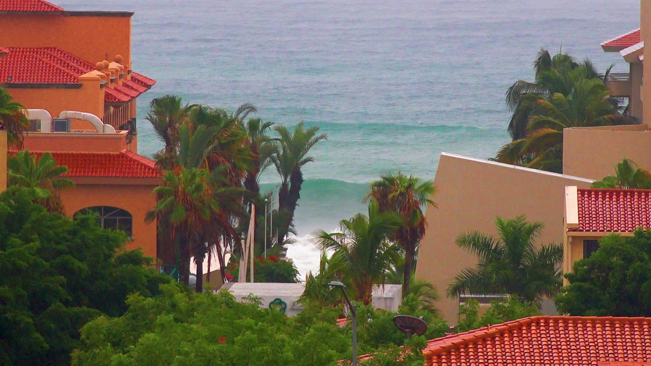 San Jose del Cabo villas with red tile roofs and palms frame a gray-blue ocean with a low line of white surf, no visible people.
