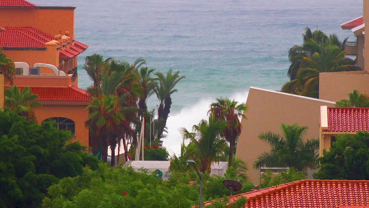 San Jose del Cabo coastal view with peach buildings, red tile roofs, palms, and white surf breaking behind them, with no visible people.