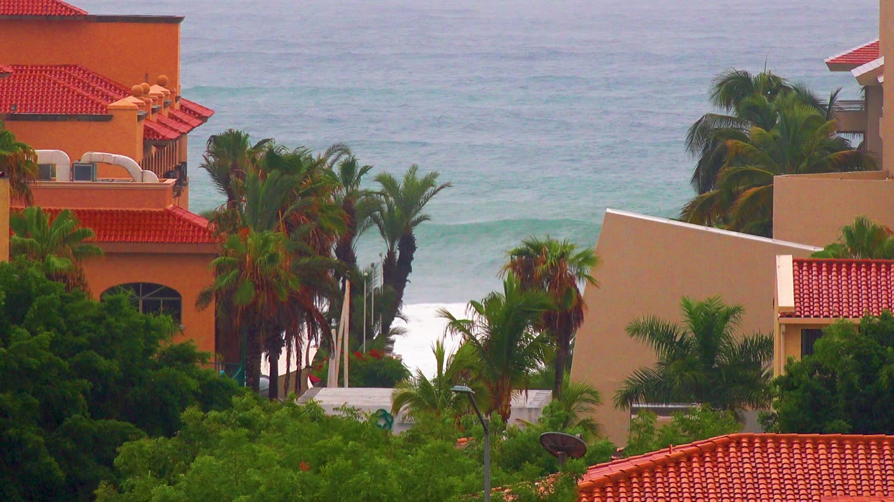 San Jose del Cabo orange stucco buildings with red tile roofs, palm trees, pale ocean water, white surf, and no visible people at the start of the clip.