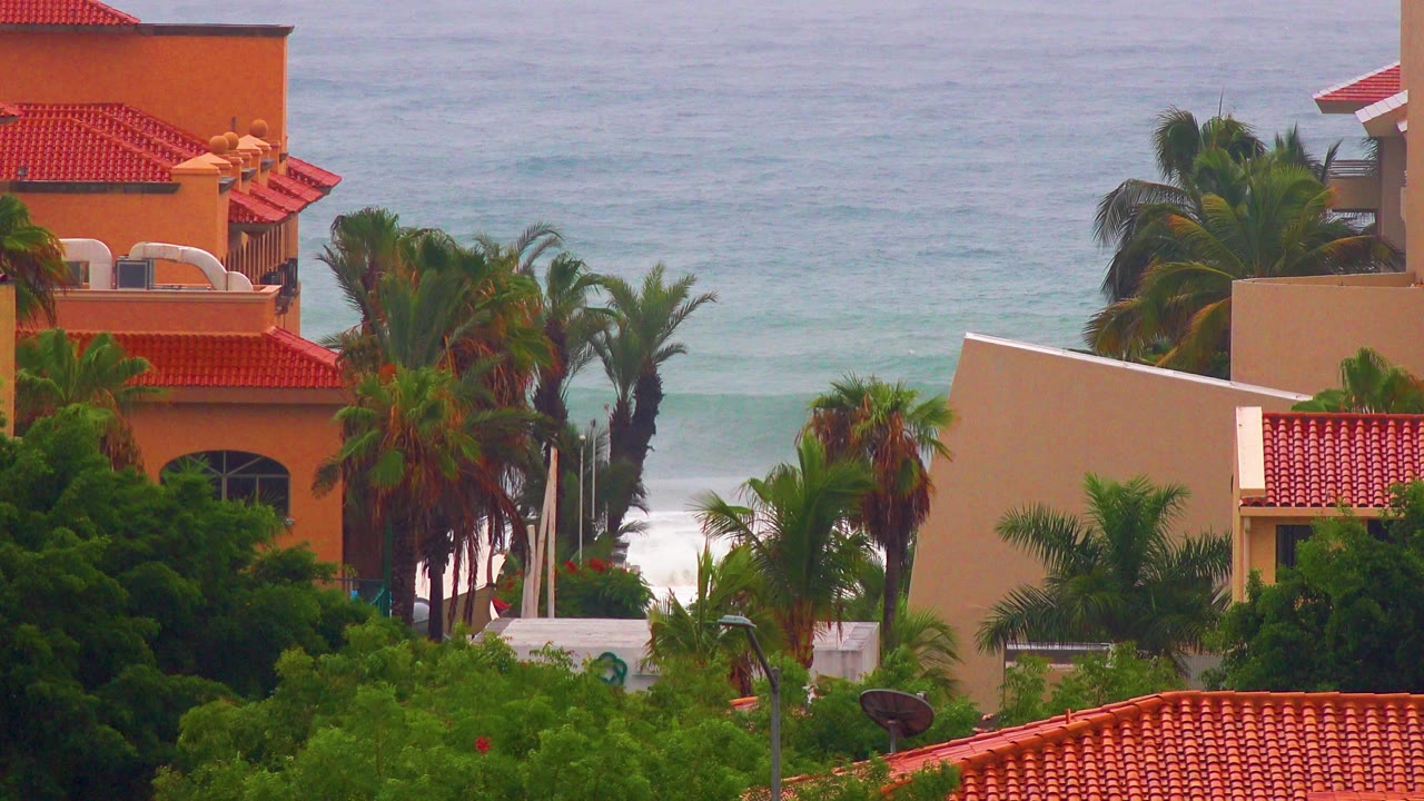 Opening frame of a San Jose del Cabo resort-front view with red tile roofs, palms, green landscaping, pale ocean water, and no visible people.