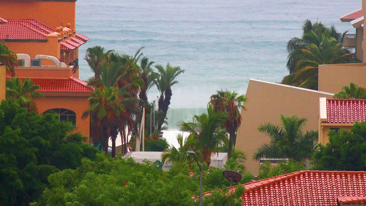 San Jose del Cabo coastal view with red tile roofs, palms, orange stucco buildings, and pale blue surf in the distance, with no visible people.
