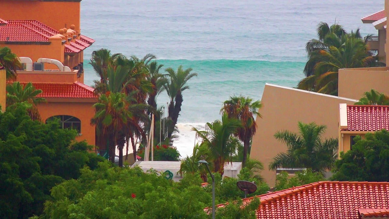 San Jose Del Cabo coastal buildings with red tile roofs and palms in front of pale blue ocean waves, no visible people.