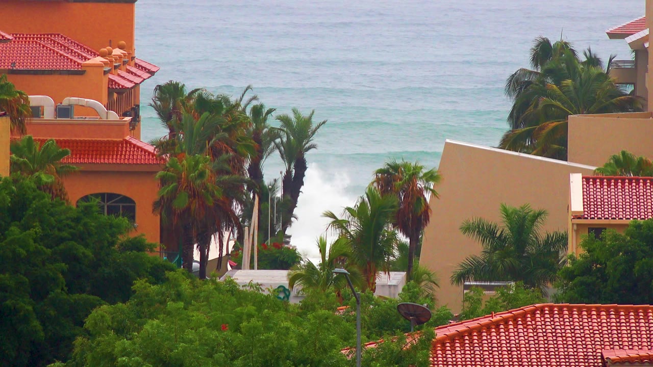 San Jose del Cabo coastal view with orange and beige building walls, red roof tiles, dense palms, and a brighter patch of white surf at left, with no visible people.