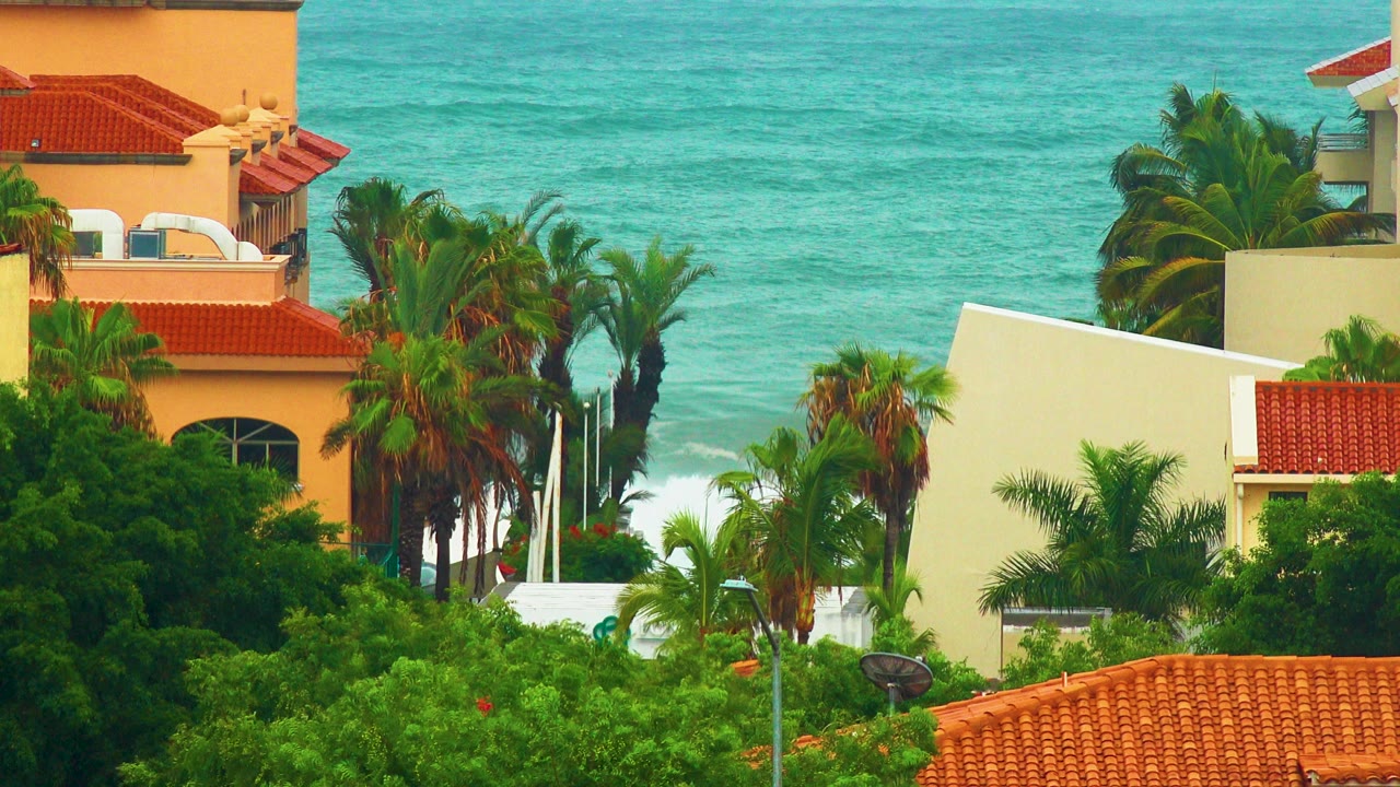 Opening frame of San Jose del Cabo beach homes with red tile roofs, palms, and blue ocean waves breaking in the background, with no visible people.