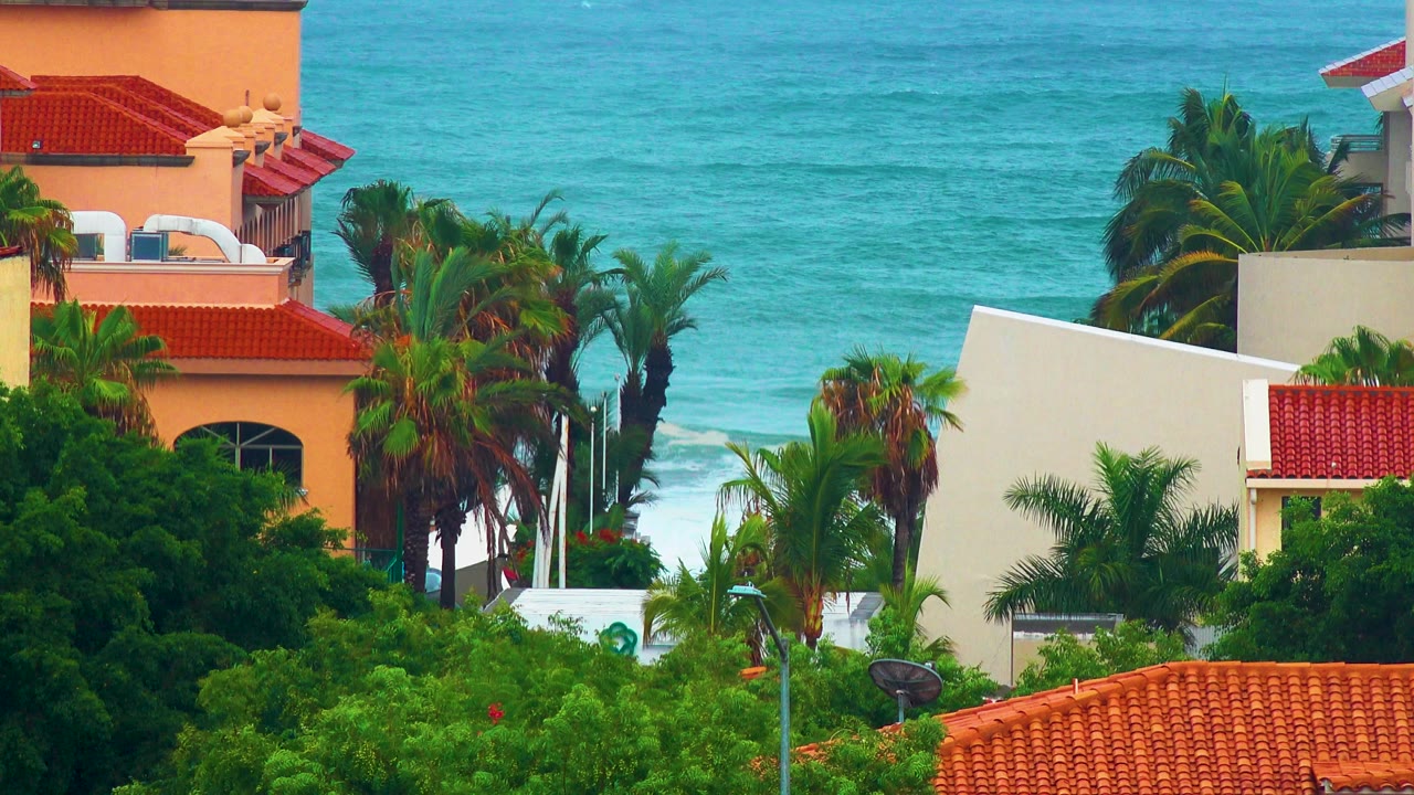 Opening frame in San Jose del Cabo showing red tile roofs, cream buildings, palms, turquoise water, white surf, and no visible people.