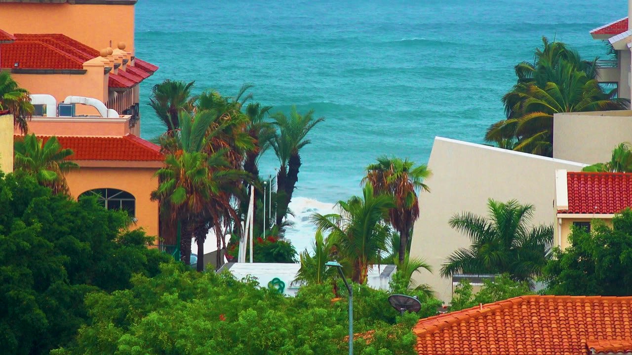 Opening view in San Jose del Cabo shows green palms, orange and cream buildings, red tile roofs, and turquoise ocean in the background with no visible people.