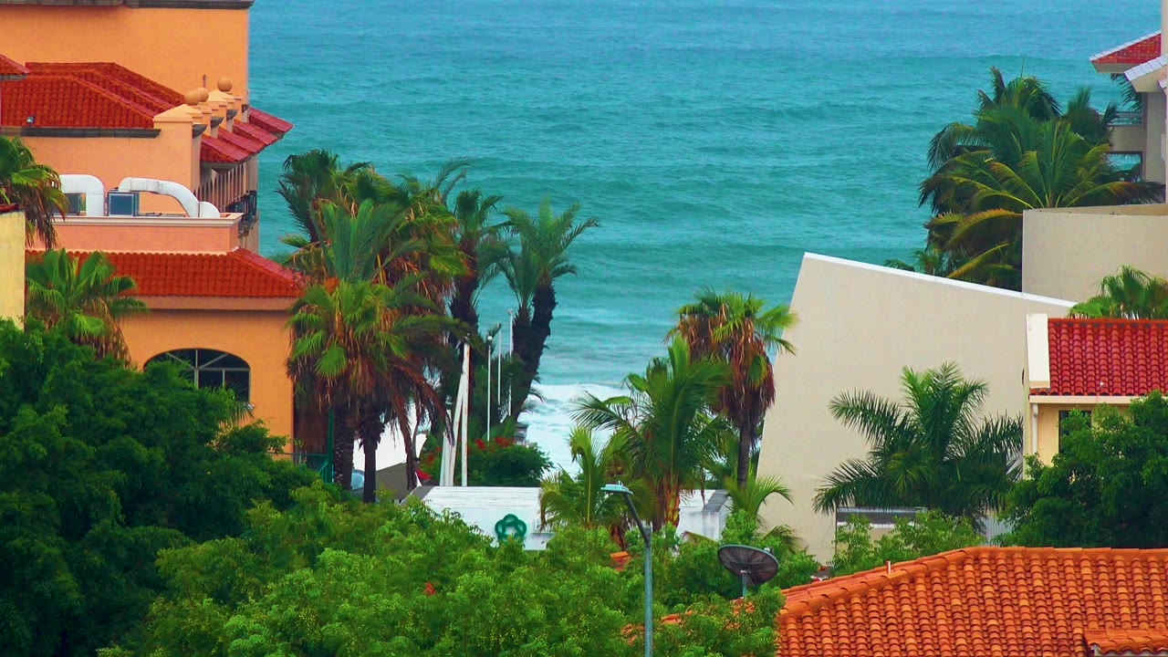San Jose del Cabo oceanfront houses with red tile roofs, green palms, and turquoise water in the background, with no visible people.