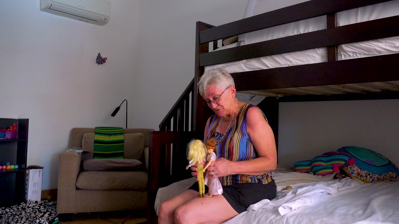 In the Los Cabos interior, the woman holds a blonde doll and a smaller doll upright while seated near white sheets and patterned pillows under the bunk bed.