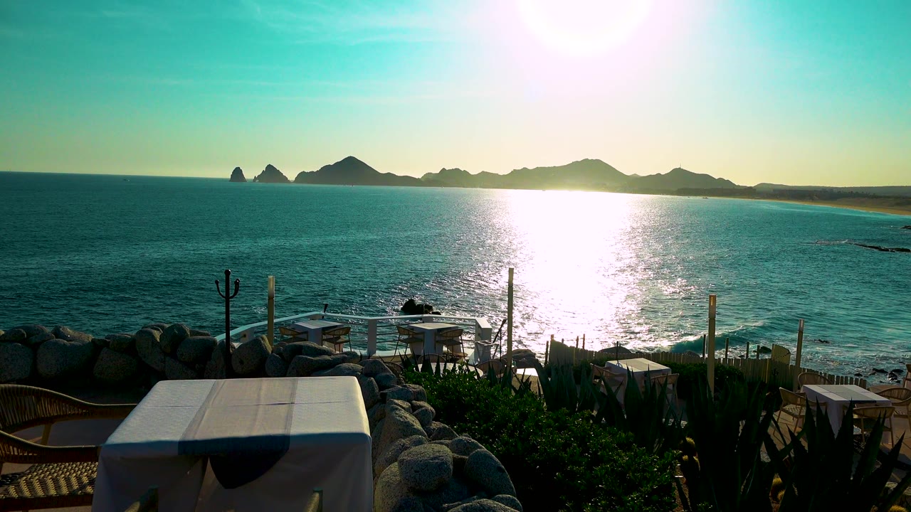 Cabo San Lucas collection view with strong glare across the sea, a dark mountain ridge on the horizon, terrace furniture and rail posts at the bottom, and no visible people.