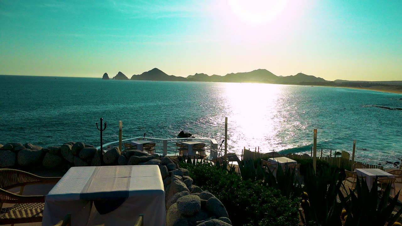 Cabo San Lucas seaside terrace view one second in, with stronger water glare near center, foreground seating and plants, distant rocky silhouettes, and no visible people.