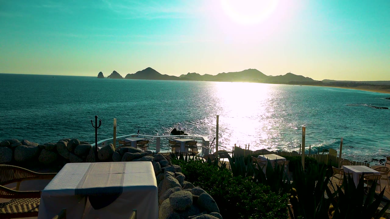 Cabo San Lucas terrace viewpoint with a white-covered table or bench in the lower frame, shimmering ocean beyond, distant land on the horizon, and no visible people.
