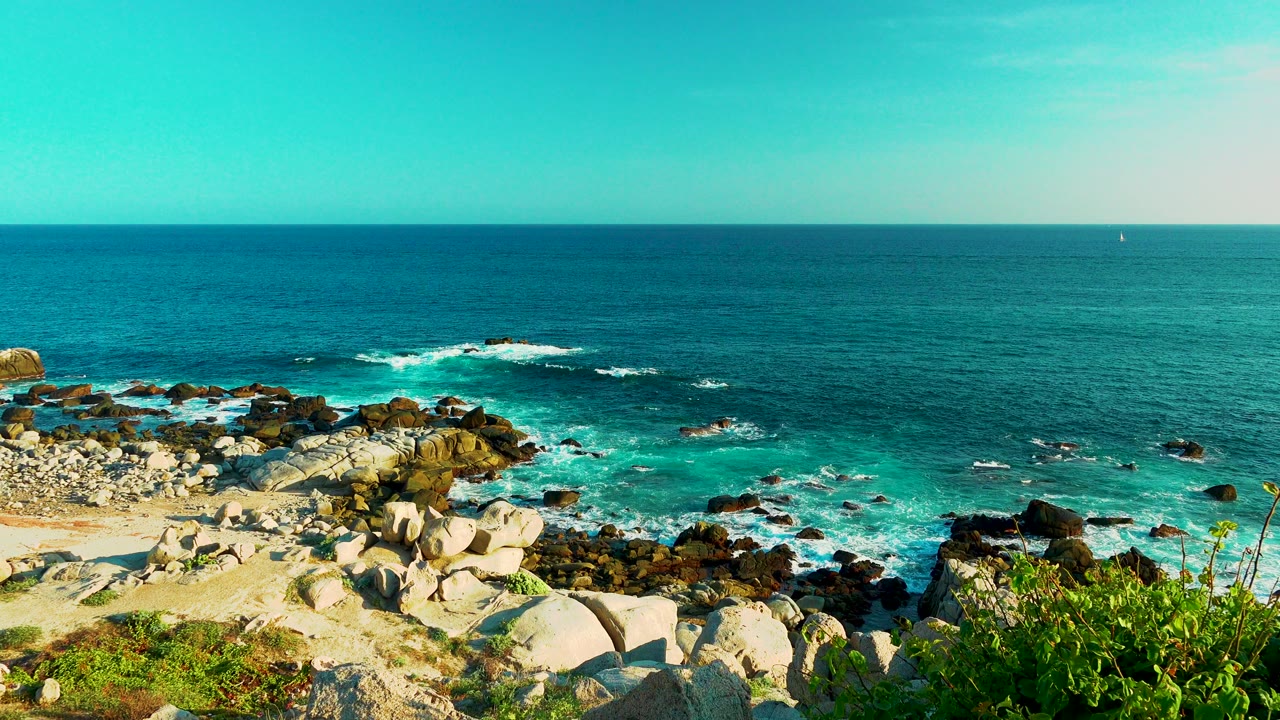 Cabo San Lucas ocean view with white waves breaking farther out around dark rocks, pale shoreline stones in the lower frame, green plants at right, and no visible people.