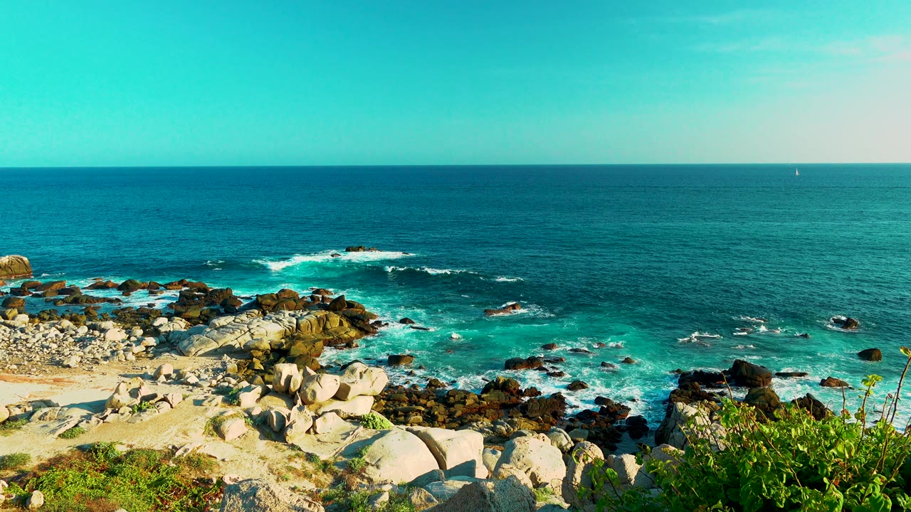 Cabo San Lucas shoreline frame with bright turquoise water near the rocks, white foam across the midground, foreground boulders and shrubs, distant horizon, and no visible people.