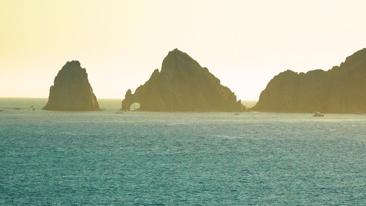 Cabo San Lucas sea cliffs remain centered across calm teal water, with an arched opening visible in one rock, tiny boats, yellow haze, and no visible people.