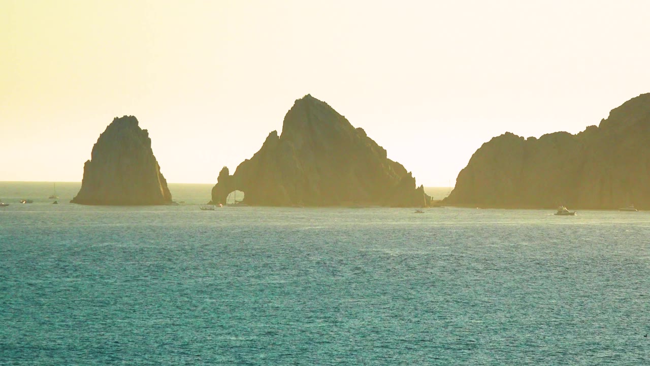 A wide Cabo San Lucas frame shows dark offshore cliffs stretching across the horizon, boats spaced along the waterline, textured teal water, and no visible people.
