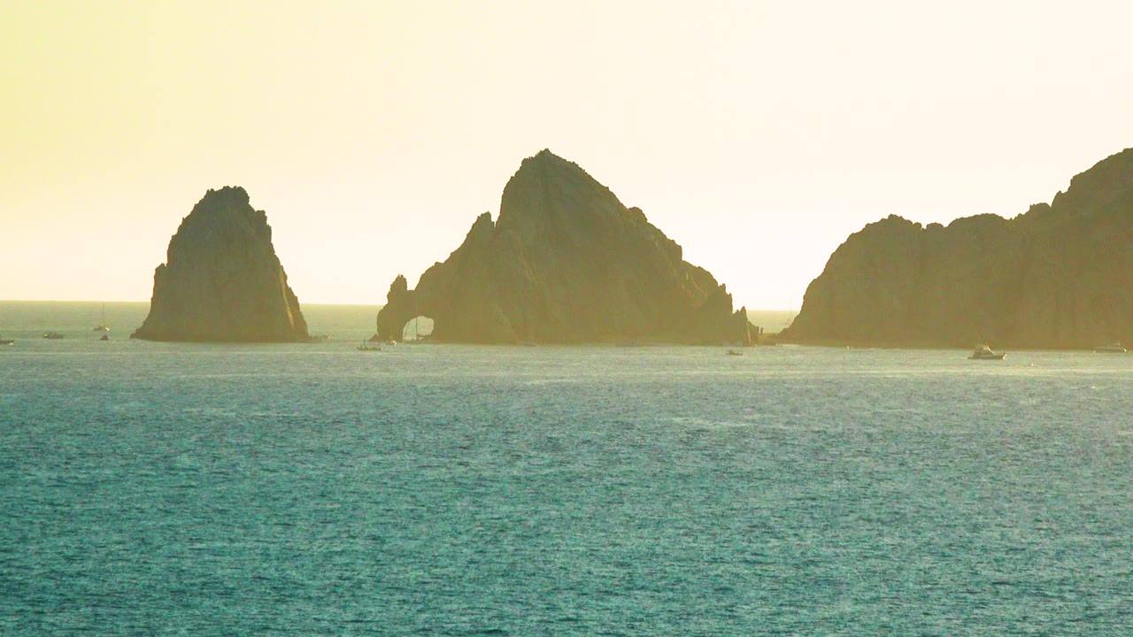 Wide Cabo San Lucas ocean view with turquoise water, dark rock formations on the horizon, small distant boats, warm haze, and no visible people.