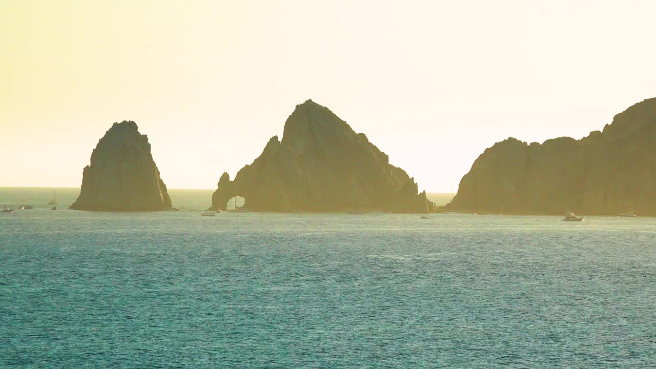 A wide Cabo San Lucas seascape shows calm water in the foreground, sea stacks across the horizon, a small arch, distant boats, and no visible people.