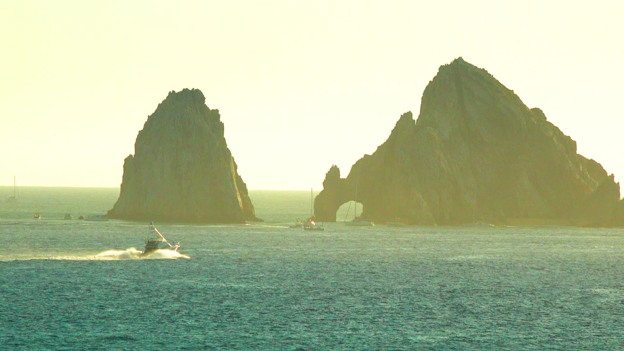 A small vessel crosses blue-green water near two large offshore rock formations in the Cabo San Lucas collection, with hazy sun and no visible people.