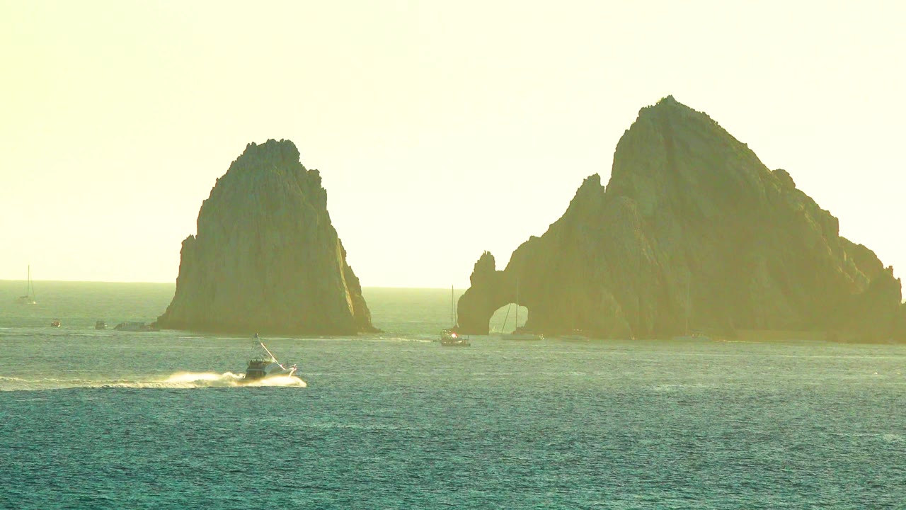 A small boat runs across the foreground water with a bright white wake below dark offshore rocks and a small arch opening, with no visible people.