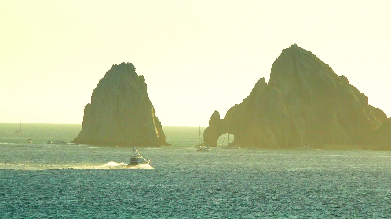 The boat sits slightly farther along the foreground, throwing spray across the ocean while the large rock mass and arch stay clear in the background, with no visible people.