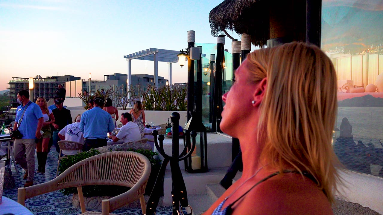 A blond woman fills the right foreground as adults sit and stand on a crowded terrace in Cabo San Lucas, Mexico, with glass panels, chairs, plants, and pale sunset sky behind them.